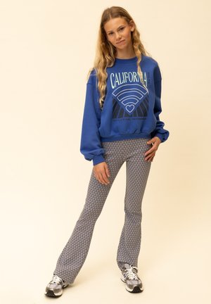 Blauwe trui met de opdruk "CALIFORNIA" en wijde broek met patroon in tinten blauw en wit, gecombineerd met grijze sneakers.