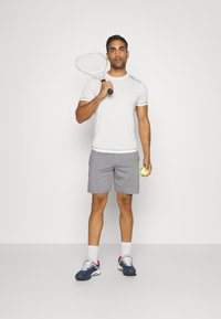 Hvit athletic t-skjorte med korte ermer, grå shorts, holder en tennisracket og ball. Joggesko med blå, hvite og røde detaljer.