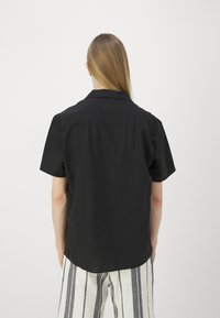 Osoba z długimi prostymi włosami, ubrana w czarną koszulkę z krótkim rękawem i białe spodnie w pionowe czarne paski, stoi tyłem na jednolitym tle.