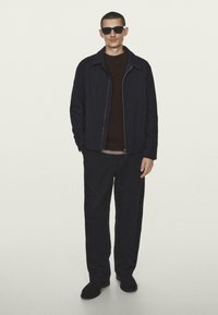 Mand iført sorte solbriller, sort jakke, mørkebrun sweater, sorte bukser og sorte sko, som står foran en ensfarvet lys baggrund.