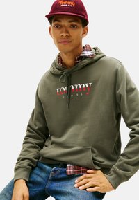 Felpa verde oliva con tasca frontale e logo bianco/rosso, indossata sopra una camicia a quadri. Un berretto bordeaux con logo completa il look.