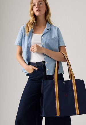 Ženska oblečena v svetlo modro kratko slečeno srajco, belo majico brez rokavov, temne jeans hlače, drži mornarsko torbo s rumenimi in maroon črtastimi ročaji.
