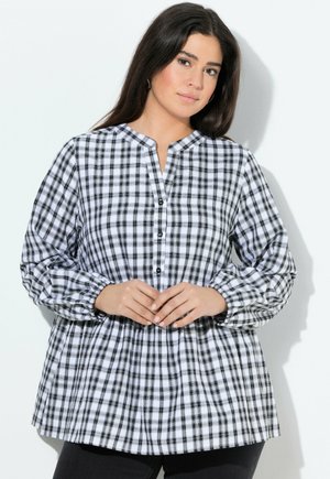 Žena v černobílé kostkované halence s dlouhým rukávem a zapínáním na knoflíky, stojící proti bílému pozadí se sepnutýma rukama.