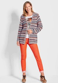 Multikleurige gebreide cardigan met strepen in marineblauw, koraal en room, gecombineerd met feloranje strakke broek en bruine platformsandalen.
