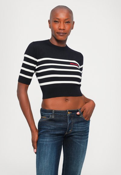 Model wearing black and white striped cropped sweater with red logo, paired with dark blue denim jeans, posing with one hand in pocket.