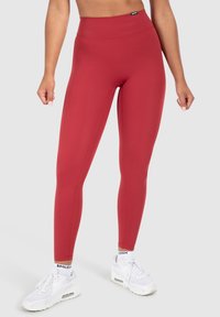 Red high-waisted leggings made of stretchy material. Smooth texture with flat seams. Paired with white athletic shoes.