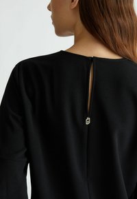 Abito nero con un design a chiave sul retro, tessuto testurizzato e un piccolo logo metallico come accentuazione alla chiusura.