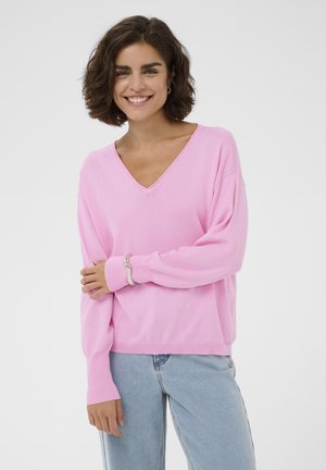 Jeune femme aux cheveux bruns courts et bouclés, portant un pull rose à col en V et un jean bleu clair, souriante, les bras croisés.