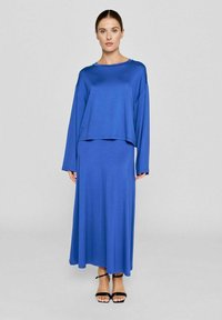 Blå, langærmet crop top parret med en matchende lang nederdel, begge lavet af glat stof. Modellen bærer sorte, stroppede sandaler.