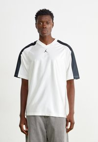 Camisa deportiva blanca con detalles negros, de manga corta y con cuello en V. Presenta un pequeño logo negro en el pecho. Textura suave, diseño casual.