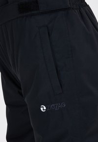 Gros plan sur un pantalon noir avec une poche latérale zippée et un logo "ZIGZAG" brodé en blanc près de la poche sur une ceinture élastique.