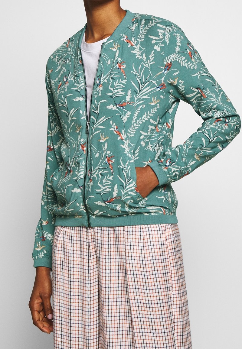 Personne portant une veste verte zippée avec un motif d’oiseau et de feuillage sur un haut blanc et une jupe à carreaux, les mains partiellement dans les poches.