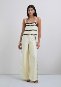Top de punto amarillo con bordes en navy con forma de concha, combinado con pantalones anchos amarillos que presentan rayas en navy a lo largo de los lados. Sandalias completan el look.