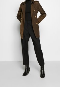 Person som bär en brun dubbelknäppt jacka, svart polotröja, svarta byxor och svarta klackiga ankelstövlar, står med händerna i fickorna.