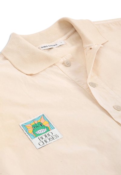 Chemise beige clair à manches courtes avec col tricoté, trois boutons à l'avant et un écusson représentant une grenouille verte souriante avec le texte "BOBO CHOSES".
