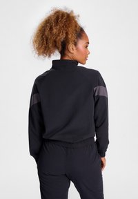 Svart idrottsjacka med hög krage, med grå axeldetaljer, elastiska manschetter och en mjuk yta för komfort och flexibilitet.