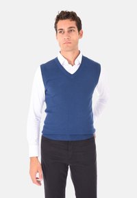 Gilet senza maniche blu lavorato a maglia con scollo a V, indossato sopra una camicia bianca con colletto. Texture liscia e silhouette aderente. Senza fantasie, risultando semplice.