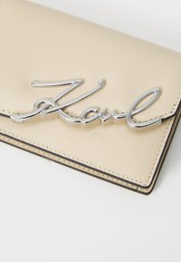 Beige leren portemonnee met een zilveren metalen "Karl" handtekeningdetail op de voorkant, met zichtbare stiksels en nette randen.