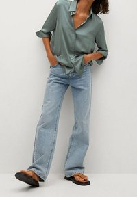 Personne portant un jean large bleu clair, une chemise verte à manches longues avec les poignets retroussés, et des sandales noires, debout les mains dans les poches.