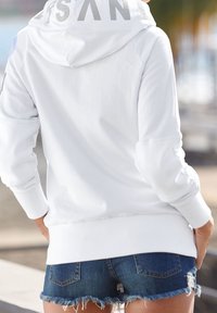 Persona che indossa un hoodie bianco con testo sul cappuccio e pantaloni di jeans strappati, in piedi all'aperto con le mani nelle tasche.