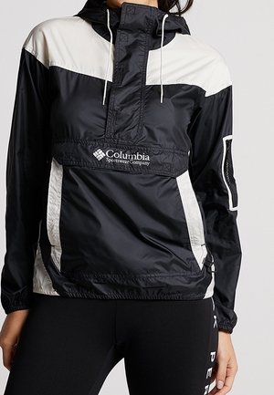 Chaqueta rompevientos Columbia en blanco y negro con capucha y bolsillo delantero, usada por una persona que está delante de un fondo liso.