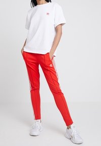 Vit kortärmad t-shirt med röd logotyp, kombinerad med röda slim-fit byxor med vita sidostråk och vita sportskor.