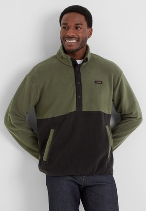 Fleece pullover in olive green with black lower section, half-zip design, button accents, and front pockets. Soft texture, casual fit.