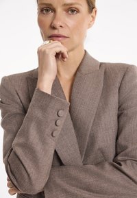Brązowa strukturalna marynarka z szalowym klapem i guzikami. Materiał wydaje się miękki i dopasowany, o lekko luźnym kroju.