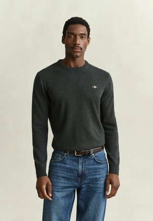 Homme portant un pull gris foncé à manches longues et un jean bleu, debout devant un fond clair uni.
