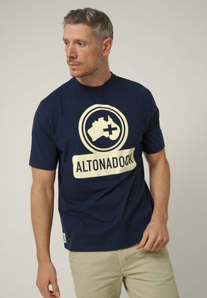 Hombre con camiseta azul marino Altonadock con logo beige y pantalones caqui, mirando hacia la izquierda contra fondo liso.