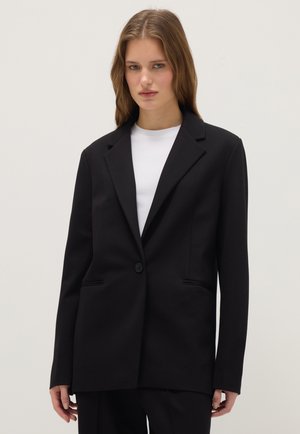 Jeune femme portant un blazer noir à un seul bouton sur une chemise blanche, debout devant un fond clair uni.