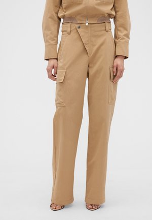 Pantalon cargo beige taille haute à jambes larges avec poches à rabat, porté avec une veste beige assortie et des talons ouverts.