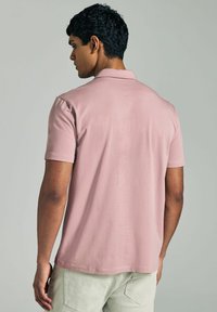 Paita polo valmistettu pehmeästä vaaleanpunaisesta kankaasta, jossa on klassinen kaulus, lyhyet hihat ja rento istuvuus, yhdistettynä vaaleisiin housuihin.