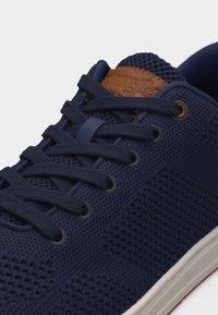 Tumesinise lõngaga tossud tumesiniste paeltega, valge tallaga ja pruuni nahast keelemärgiga, rõhutades esikülje ja külje detaile.