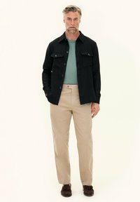 Black button-up shirt jacket with two chest pockets, worn over a light green shirt, paired with beige trousers and brown shoes.
