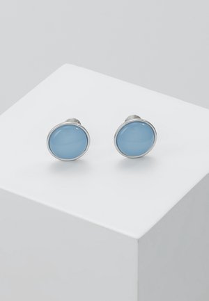 Paire de boutons de manchette en argent avec incrustations de pierres rondes, lisses et bleu clair, reposant sur un socle cubique blanc.