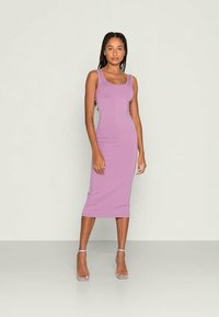 Femme debout portant une robe midi sans manches ajustée lilas et des escarpins à sangle cheville lilas contre un fond clair uni.