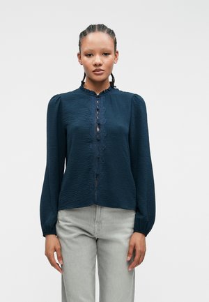 Ναυτικό μπλε μπλούζα με υφή, ραμμένο γιακά και κουμπιά μπροστά, διακοσμημένα με δαντέλα. Μακριά μανίκια με συγκεντρωμένα μανίκια, συνδυασμένα με γκρίζα τζιν.