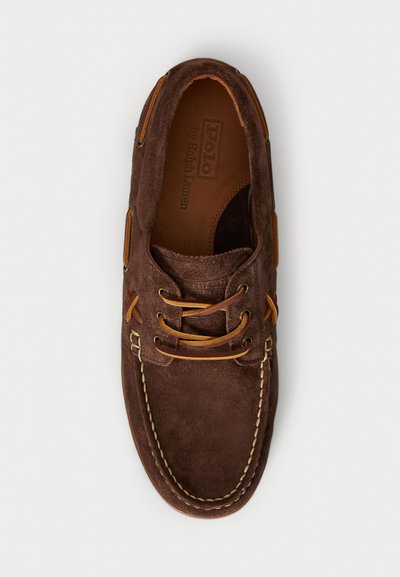 Zapato tipo mocasín de ante marrón con acentos de piel, costuras en contraste y cordones circulares. El interior presenta un logo y un forro suave.