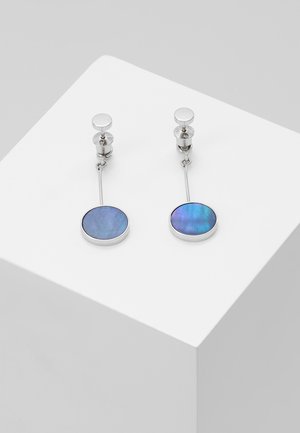 Boucles d'oreilles en argent avec pendentifs ovales irisés bleus, présentées sur un cube blanc sur un fond gris.