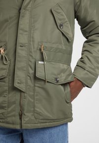 Olivgrüne isolierte Jacke mit Toggle-Details, großen Fronttaschen und Reißverschluss. Glatte Textur mit Knopfakzenten.