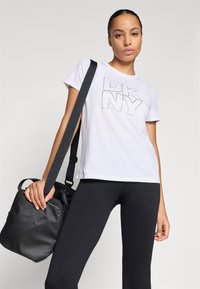 Balta T-kreklu ar "DKNY" logotipu teksturētā, kontūrētā dizainā. Kombinēts ar melnām teksturētām legingām un melnu pleca somu.