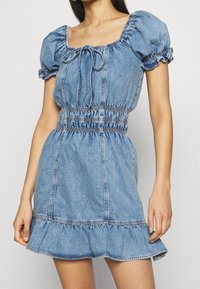 Denimmekko, jossa on lyhyet puff-hihat, neliön muotoinen pääntie ja naru-tyyliset yksityiskohdat vyötäröllä. Ominaisuuksiin kuuluu röyhelöhelma ja vaaleansininen rakenne.