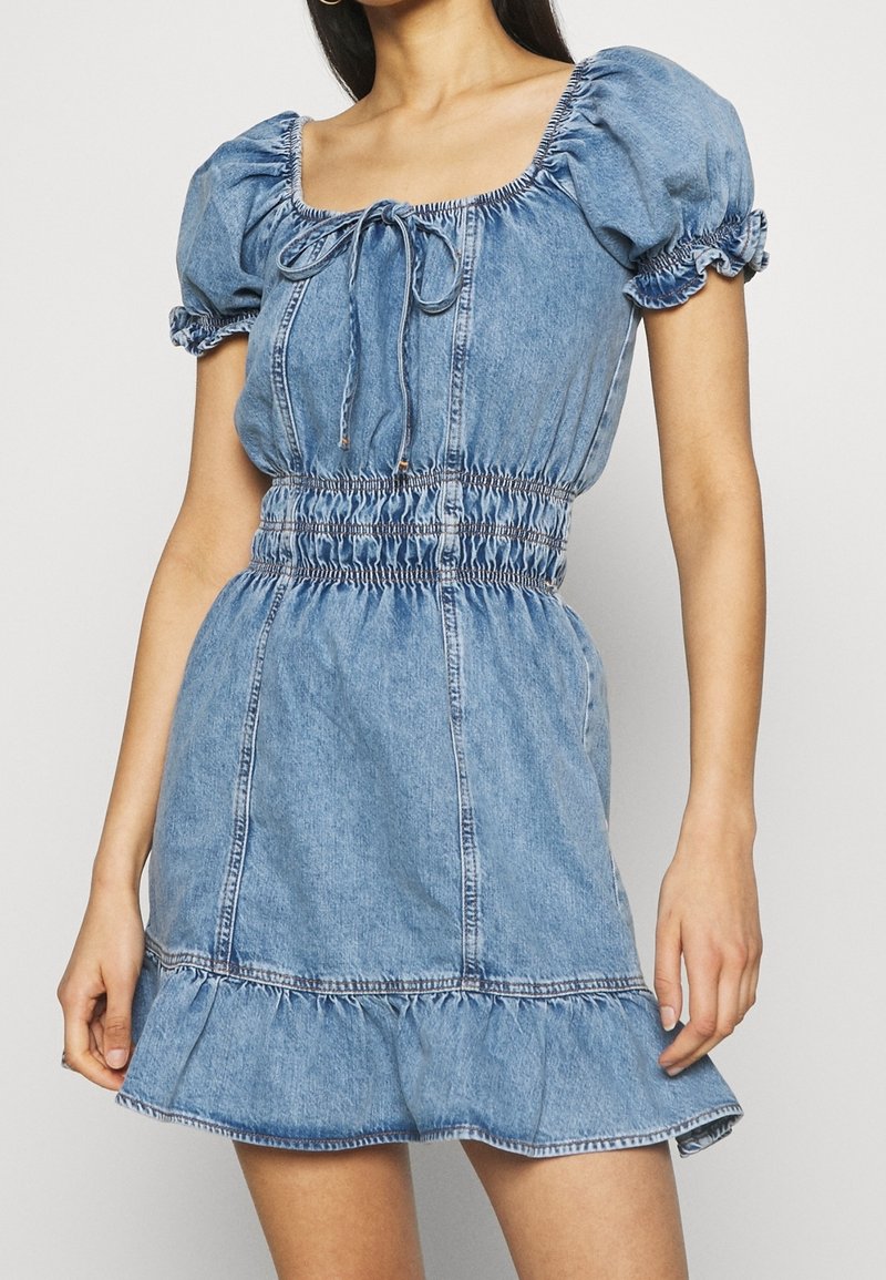 Denimmekko, jossa on lyhyet puff-hihat, neliön muotoinen pääntie ja naru-tyyliset yksityiskohdat vyötäröllä. Ominaisuuksiin kuuluu röyhelöhelma ja vaaleansininen rakenne.