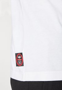 White cotton t-shirt with a black and red logo patch featuring three stars, positioned at the bottom left seam of the shirt.