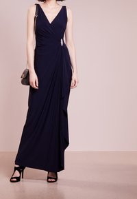 Navy evening gown with a deep V-neck, draped design, and an embellished waist detail, paired with black strappy heels and a small bag.