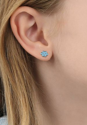 Primo piano dell'orecchio di una persona che indossa un piccolo orecchino a perno a forma di nuvola sorridente blu su pelle chiara e capelli biondi.