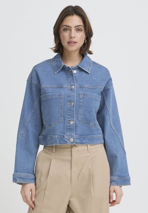 Kobieta o krótkich brązowych włosach, ubrana w niebieską krótka kurtkę dżinsową z dużymi guzikami i beżowe, plisowane spodnie z wysokim stanem.