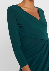 Sjögrön klänning med V-ringning, långa ärmar och draperad tygdetalj. Tillverkad av ett slätt, elastiskt material med en figurnära silhuett.