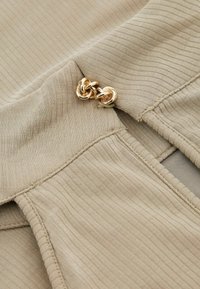 Beige ribattu kangas, jossa on kultainen metallinen solkiyksityiskohta. Kankaan pinta on sileä, ja siinä on hienosti sijoitettuja pystysuoria viivoja sekä ommeltuja reunoja.